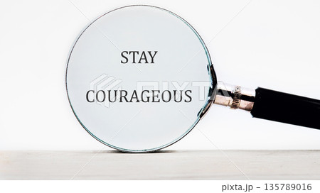 A magnifying glass rests on a light colored surface, revealing the bold words STAY COURAGEOUS beneath its lens. The background is a stark white 135789016