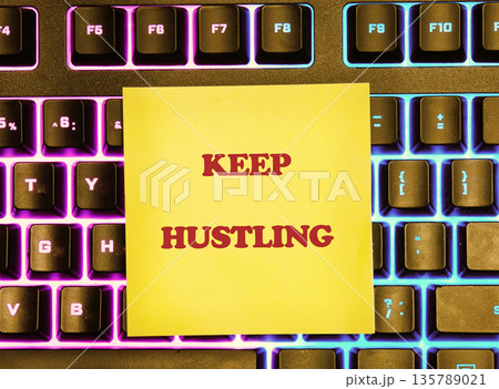 Yellow sticky note with KEEP HUSTLING in red bold text rests on a black computer keyboard. The keys glow with bright purple and blue backlighting, creating an inspiring work scene 135789021