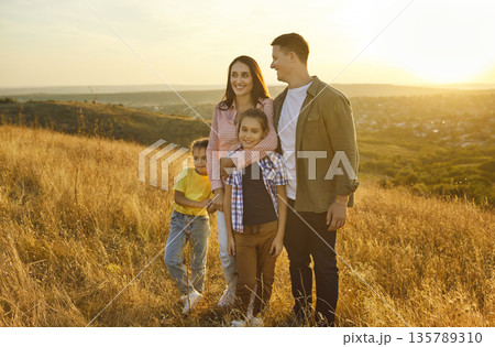 Happy family standing together at sunset, parents hug children, enjoy golden light Happy family standing together at sunset, parents hug children, enjoy golden light 135789310