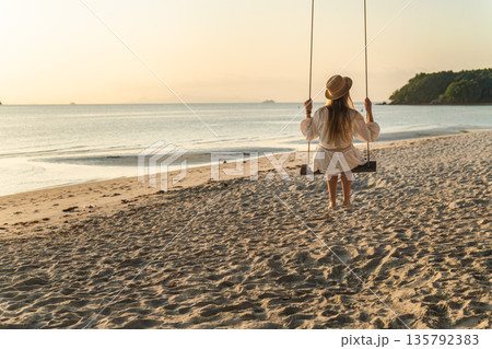 Young woman sitting on wooden swing on sandy tropical beach at sunset and sunrise light facing calm sea on Koh Samui Thailand Young woman sitting on wooden swing on sandy tropical beach at sunset and sunrise light facing calm sea on Koh Samui Thailand 135792383