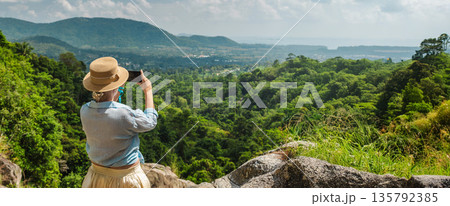 Young woman in straw hat photographing tropical mountain valley with smartphone from rocky viewpoint, lush green forest, distant hills and wide copy space for travel concept Young woman in straw hat photographing tropical mountain valley with smartphone from rocky viewpoint, lush green forest, distant hills and wide copy space for travel concept 135792385