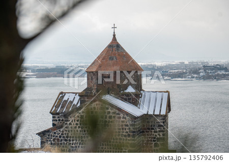 Stone church of Sevanavank Monastery stands above snowy shore of Lake Sevan in Armenia, dark volcanic masonry contrasts with pale winter sky 135792406