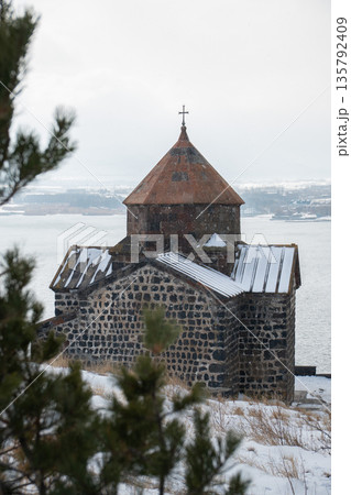 Stone church of Sevanavank Monastery stands above snowy shore of Lake Sevan in Armenia, dark volcanic masonry contrasts with pale winter sky 135792409