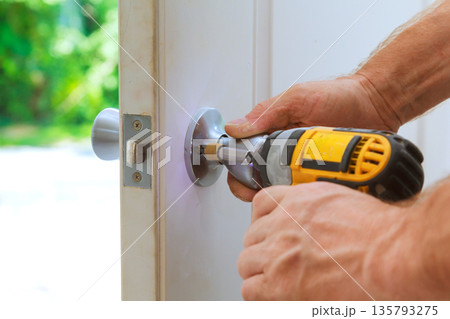 Carpenter is using drill to fix door handle onto door in home setting 135793275