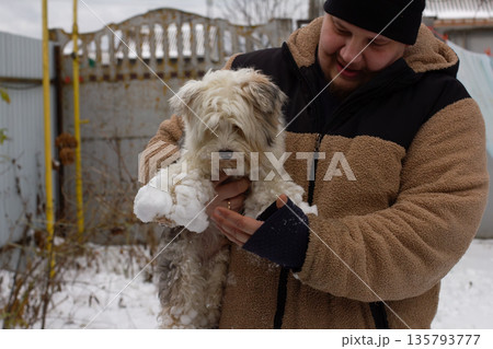 A man smiles, holds his fluffy white dog with snow on his fur and paws, sharing playful, affectionate winter moments outdoors in warm clothes and a hat. A large amount of snow stuck to the dog's paws. 135793777