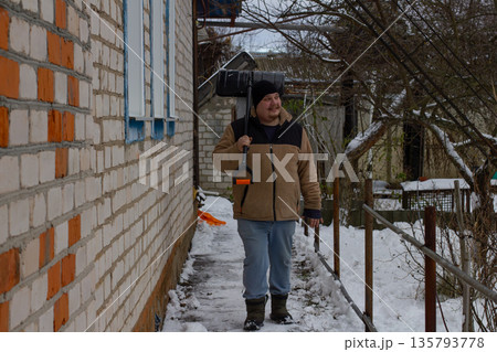 A man carries a black snow shovel on his shoulder and walks along a narrow snow-covered path along the wall of a house in winter, preparing to clear snow. A man goes to clear the snow. A man carries a black snow shovel on his shoulder and walks along a narrow snow-covered path along the wall of a house in winter, preparing to clear snow. A man goes to clear the snow. 135793778
