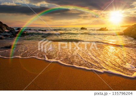 beautiful sunset over empty sandy beach at the sea. white foam rolling on to the shore near boulder in evening light with rainbow. perfect summer vacation concept. awesome weekend. low angle view 135796204
