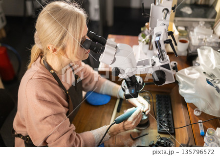 Woman jeweler crafting with precision using microscope Woman jeweler crafting with precision using microscope 135796572
