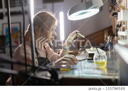 Woman soldering metal artwork in jewelry workshop Woman soldering metal artwork in jewelry workshop 135796589