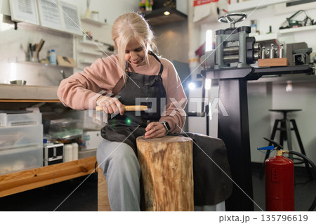 Woman jeweler artisan crafting metal with hammer in workshop Woman jeweler artisan crafting metal with hammer in workshop 135796619