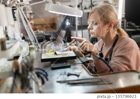 Woman jeweler soldering metal piece in workshop Woman jeweler soldering metal piece in workshop 135796623