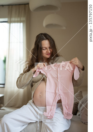 Pregnant woman holding pink baby clothes sitting on sofa 135797150