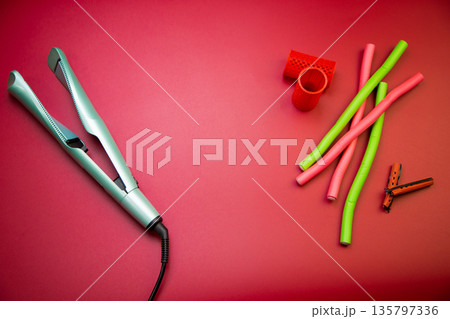 Top view of a green hair straightener with a swirling ceramic canvas, next to which there are curlers of different shapes and sizes on a pink background with space for text. The concept of styling Top view of a green hair straightener with a swirling ceramic canvas, next to which there are curlers of different shapes and sizes on a pink background with space for text. The concept of styling 135797336