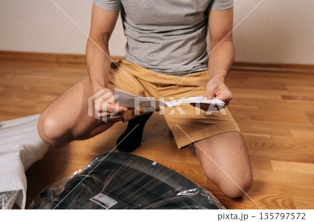 Cropped shot of man following written instructions to assemble new flat-pack furniture, focusing on steps to build office chair on te hardwood floor, common home improvement and diy task. 135797572