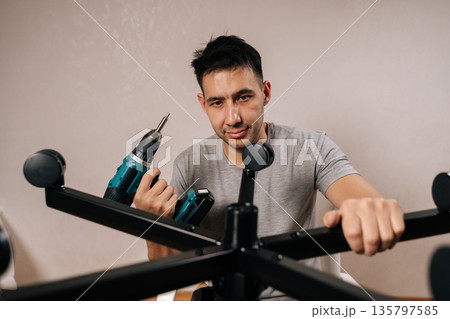 Portrait of contractor male assembling new office chair indoors, using electric drill to secure parts, focused diy furniture project showing tools, skill and home workspace setup. 135797585