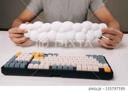 Person holding cloud-shaped ergonomic wrist rest for keyboard, providing comfortable support and wrist pain prevention during long hours of office work or gaming, promoting healthy posture, close-up. Person holding cloud-shaped ergonomic wrist rest for keyboard, providing comfortable support and wrist pain prevention during long hours of office work or gaming, promoting healthy posture, close-up. 135797589