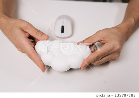 Person holding cloud-shaped wrist rest for computer mouse, providing ergonomic support and comfort while working on home office desk, preventing carpal tunnel syndrome and repetitive strain injury. Person holding cloud-shaped wrist rest for computer mouse, providing ergonomic support and comfort while working on home office desk, preventing carpal tunnel syndrome and repetitive strain injury. 135797595