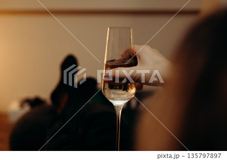 A hand holding a flute glass filled with sparkling wine or champagne. The image focuses on the drink and glassware against a warmly lit interior A hand holding a flute glass filled with sparkling wine or champagne. The image focuses on the drink and glassware against a warmly lit interior 135797897