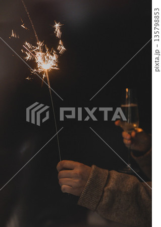 A person holding a burning sparkler and a glass of champagne in a dark, cozy atmosphere. Festive New Year or Christmas celebration, holiday magic A person holding a burning sparkler and a glass of champagne in a dark, cozy atmosphere. Festive New Year or Christmas celebration, holiday magic 135798853
