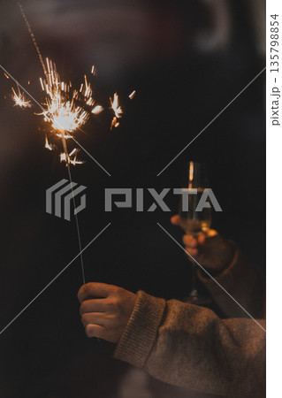 A person holding a burning sparkler and a glass of champagne in a dark, cozy atmosphere. Festive New Year or Christmas celebration, holiday magic A person holding a burning sparkler and a glass of champagne in a dark, cozy atmosphere. Festive New Year or Christmas celebration, holiday magic 135798854