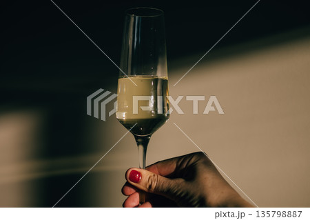 Female hand holding champagne glass with red manicure in dramatic light and shadow. Festive toast, celebration, holiday greeting, elegant lifestyle. 135798887