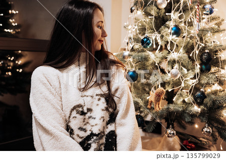 A woman in a fluffy sweater looking at the softly lit Christmas tree in the evening. This authentic, candid moment is perfect for lifestyle content focused on home celebrations and the festive spirit. 135799029