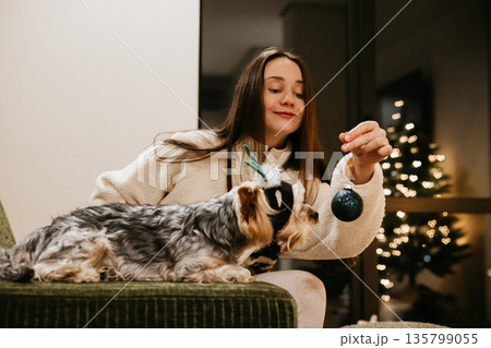Playful Christmas lifestyle scene at home - a young woman playing with a lovable small yorkie dog in cozy modern interior with festive lights, pet-friendly holiday trend and warm winter mood scene. Playful Christmas lifestyle scene at home - a young woman playing with a lovable small yorkie dog in cozy modern interior with festive lights, pet-friendly holiday trend and warm winter mood scene. 135799055