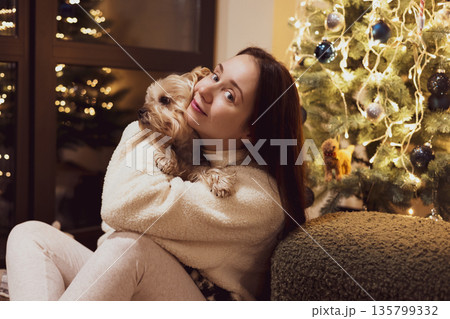 Festive Christmas moment at home - a young white woman sharing a cozy, joyful evening with a small dog Yorkshire terrier near a decorated Christmas tree, warm lights, hygge mood and holiday lifestyle. Festive Christmas moment at home - a young white woman sharing a cozy, joyful evening with a small dog Yorkshire terrier near a decorated Christmas tree, warm lights, hygge mood and holiday lifestyle. 135799332