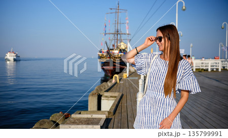 Young woman standing on pier and adjusting sunglasses by the sea Young woman standing on pier and adjusting sunglasses by the sea 135799991