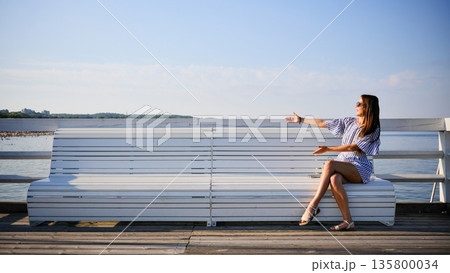 Young woman pointing at sea while sitting on bench on summer day 135800034