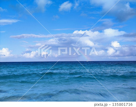 Ocean view under a bright blue sky with scattered white clouds. A rainbow is visible on the horizon, adding a touch of color between the sea and sky 135801716