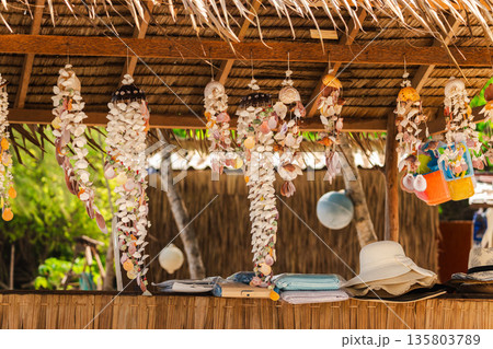 Tropical beach hut display with hanging seashell wind chimes, straw hats and folded towels under thatched roof, warm sunlight Tropical beach hut display with hanging seashell wind chimes, straw hats and folded towels under thatched roof, warm sunlight 135803789
