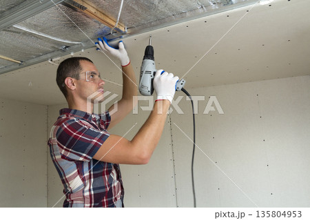Young man in goggles fixing drywall suspended ceiling to metal frame using electrical screwdriver on ceiling insulated with shiny aluminum foil. Renovation, construction, do it yourself concept. Young man in goggles fixing drywall suspended ceiling to metal frame using electrical screwdriver on ceiling insulated with shiny aluminum foil. Renovation, construction, do it yourself concept. 135804953