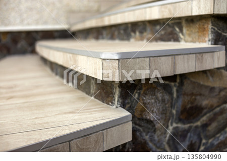 Side view of lit by sun new staircase with light tiles and decorated with natural stones building wall on blurred background. Professionally done work concept. Side view of lit by sun new staircase with light tiles and decorated with natural stones building wall on blurred background. Professionally done work concept. 135804990