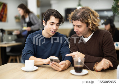 Happy guy meeting with father in cafe and drinks delicious coffee, looking at photos on mobile phone 135805935