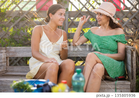 European young woman in straw hat is sitting on wooden bench and chatting sweetly with her sister 135805936