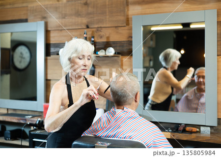 Mature woman master completes creation of senior male clients image with help of scissors 135805984