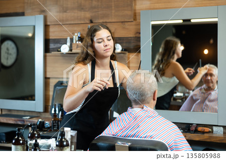 Girl master hairdresser completes creation of senior male clients image with help of scissors. 135805988