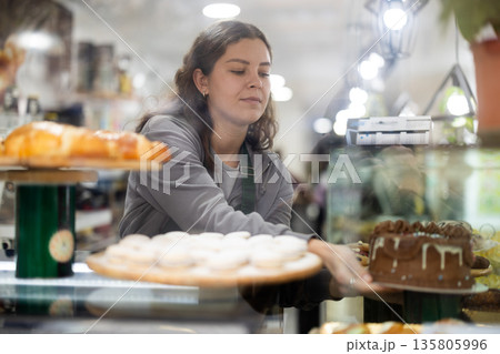Positive young woman seller putting fresh pastries on plate from counter in cafe bakery 135805996