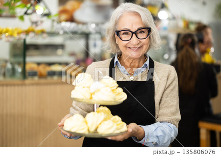 Elderly female waitress in apron invites to coffee shop and holding tray of fresh, delicious baked goods in hands 135806076