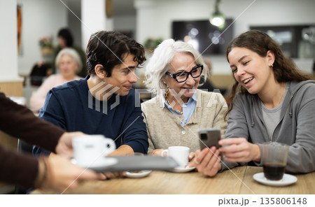 Positive elderly woman meeting and chatting with family and young couple showing photos on phone in cozy cafeteria Positive elderly woman meeting and chatting with family and young couple showing photos on phone in cozy cafeteria 135806148