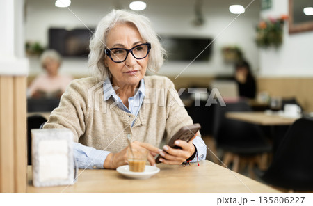 European elderly woman drinking fresh coffee at table in cafe and looking at mobile phone 135806227