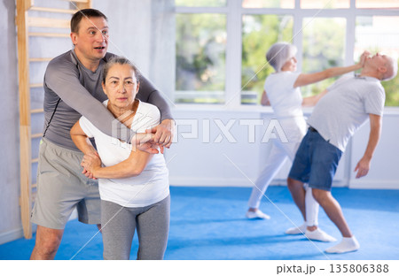 Trainer and mature woman train painful hold on self-defense course in sport gym Trainer and mature woman train painful hold on self-defense course in sport gym 135806388