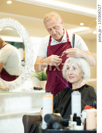 Professional male hairdresser cuts hair of elderly woman at beauty salon 135806527