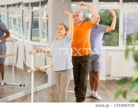 Ordinary elderly people stand in third position near ballet barre during group training in dance studio Ordinary elderly people stand in third position near ballet barre during group training in dance studio 135806559
