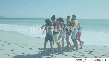 Linking arms seven friends wearing casual attire walking along beach shore, with footprints in sand 135806921