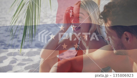 Laughing couple wearing bikini top and swim trunks on white sand under palm frond, tropical beach Laughing couple wearing bikini top and swim trunks on white sand under palm frond, tropical beach 135806923