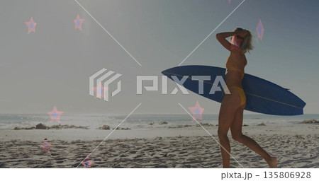 Walking surfer in yellow bikini carrying blue surfboard on beach with star lens flare, copy space Walking surfer in yellow bikini carrying blue surfboard on beach with star lens flare, copy space 135806928