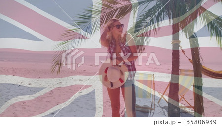 Standing in checkered shirt, shorts on sandy beach, with straw hat, palm tree, Union Jack flag 135806939