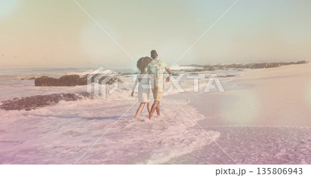 Strolling African American couple arm-in-arm into surf at sandy beach, with waves and dark rocks 135806943